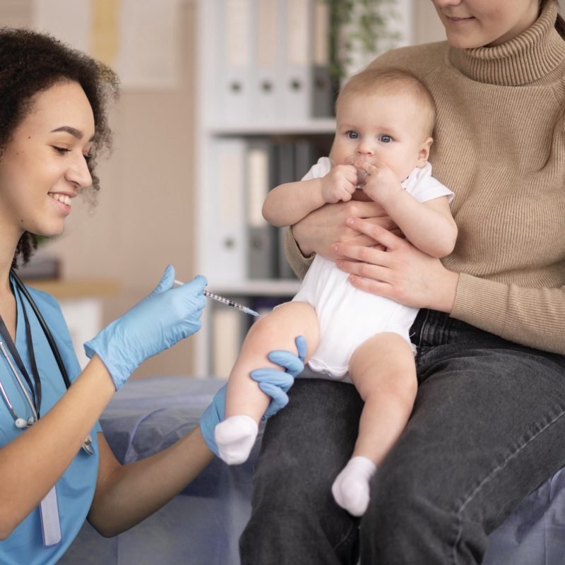 Een zorgverlener met blauwe handschoenen dient een vaccin toe aan een baby die op schoot zit bij een volwassene. De baby kijkt omhoog en de zorgverlener lacht vriendelijk.